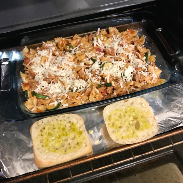 ingredients in pan, topped with cheese and garlic bread on side, going into oven